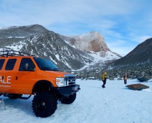 An orange expedition vehicle is parked on snowy ground in a mountainous, rocky landscape in Antarctica. Two people wearing winter gear stand nearby, surrounded by snow, rocks, and distant peaks under a partly cloudy sky.