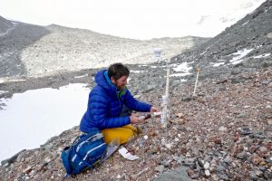 A person in a blue jacket kneels on rocky ground in snowy, mountainous Antarctica, adjusting scientific equipment attached to a pole. A blue backpack and tools from Antarctic Logistics and Expeditions are nearby.