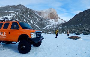 An orange expedition vehicle from Antarctic Logistics and Expeditions is parked on snow in a rocky, mountainous landscape in Antarctica. Two people in winter clothing walk nearby, surrounded by rugged peaks under a partly cloudy sky.