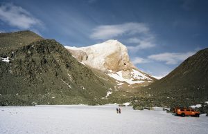 Two people stand on a snow-covered plain near a bright orange Antarctic Logistics and Expeditions vehicle, surrounded by rocky mountains and a tall, snow-capped peak under a blue sky with wispy clouds in Antarctica.