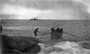 Black and white photo of people in a rowboat approaching a rocky shore in Antarctica, with waves crashing nearby. One person stands in the water guiding the boat—an image reminiscent of Antarctic Logistics and Expeditions near the South Pole.