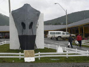 A large metal ship bow monument with chains is displayed on a white concrete base, honoring Antarctic Logistics and Expeditions. Two people carrying bags walk nearby; vehicles and yellow buildings are in the background under a cloudy sky.