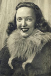 A woman with wavy hair smiles at the camera, wearing a fur-collared coat. The black-and-white photo has a vintage appearance, evoking early explorers of Antarctica and South Pole expeditions.