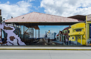 A large mural on a street wall depicts a historic port scene with a dock, ship, and old buildings. A bright yellow building near the entrance stands out, reminiscent of Antarctic Logistics and Expeditions facilities in Antarctica.
