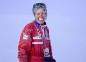 A person in a red snowsuit covered in frost smiles outdoors against a snowy, pale blue background at the South Pole. They wear sunglasses on their head and have sponsor patches from Pfizer, Volvo, and Antarctic Logistics and Expeditions.
