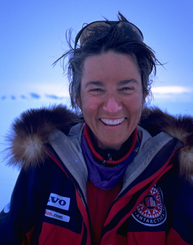 A woman in a heavy, fur-trimmed parka with Antarctic Logistics and Expeditions patches smiles outdoors against a snowy, icy South Pole backdrop. Sunglasses rest on her head and her hair is windswept.
