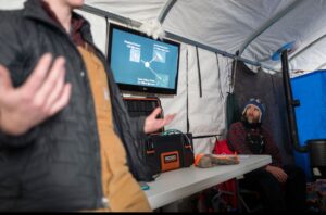A person gestures while presenting to another seated person inside a tent at the South Pole. A screen displays scientific data behind them, and tools and equipment are on the table. The seated listener, in a winter hat and overalls, is focused on Antarctic research.