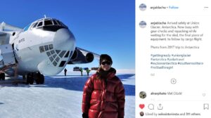 A person in a red jacket and sunglasses smiles, standing on snow near a large cargo plane at Union Glacier, Antarctica. Other people and equipment from Antarctic Logistics and Expeditions are visible under the clear blue sky.