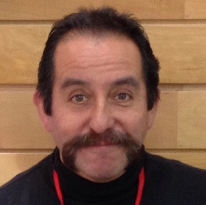 A man with dark hair and a thick mustache is smiling slightly. He is wearing a black top and a red lanyard, standing in front of a wooden background, perhaps recalling his adventures with Antarctic Logistics and Expeditions near the South Pole.