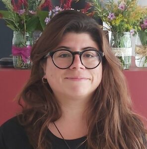 A person with long brown hair and black glasses is smiling softly. They are standing in front of a red surface at an Antarctic Logistics and Expeditions base, with several mason jars filled with colorful flowers in the background.