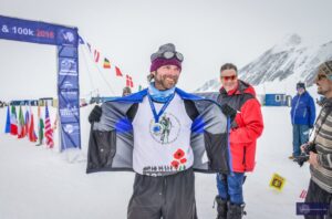 A smiling man in winter gear proudly opens his jacket to show his shirt and medals at a snowy race finish line in Antarctica, with Antarctic Logistics and Expeditions flags, mountains, and others in cold-weather clothing behind him.