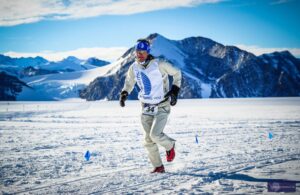 A runner in white winter gear with a race bib numbered 34 and sunglasses jogs across an Antarctic landscape marked by blue flags. Snow-covered mountains and the clear sky highlight the striking beauty of South Pole Antarctica.