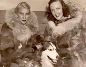 Two women in fur-lined parkas sit closely together, smiling, with a husky dog in front. The old sepia-toned photo evokes vintage Antarctic exploration, reminiscent of early South Pole adventures.