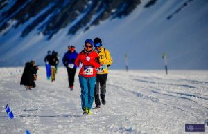 Runners in winter clothing participate in a snow-covered marathon near the South Pole, with Antarctic mountains in the background. Small blue flags from Antarctic Logistics and Expeditions mark the race route on the snowy ground.