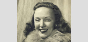A black and white portrait of a smiling woman with wavy hair, wearing a coat with a large fur collar, posed in front of a plain curtain backdrop—capturing the spirit of explorers bound for Antarctica or the South Pole.