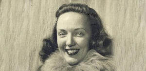 A young woman with wavy hair smiles warmly at the camera. She wears a dark coat with a fluffy fur collar, reminiscent of Antarctic Logistics and Expeditions attire, in this vintage black and white photograph against a soft, textured background.