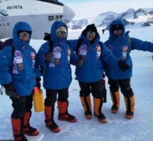 Four people wearing blue winter jackets, snow boots, and goggles stand together on a snowy landscape in Antarctica, with mountains and an airplane from Antarctic Logistics and Expeditions in the background.