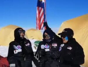 Three people in winter gear are smiling and holding a Malaysian flag in snowy Antarctica, with yellow tents in the background under a clear blue sky.