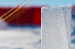 A close-up of a rectangular block of ice standing upright on snow, sparkling in the sunlight. The background is blurred with red and yellow elements, evoking the vibrant hues often seen at South Pole Antarctic Logistics and Expeditions camps.
