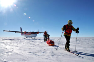 Three people in winter gear ski across a snowy, flat Antarctic landscape, pulling a sled with supplies. A small airplane from Antarctic Logistics and Expeditions is parked on the snow in the background under a clear blue sky.