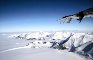 A snowy mountain range and vast white landscape in Antarctica viewed from an airplane, with part of the plane wing visible in the upper right corner under a clear blue sky—an iconic scene on Antarctic Logistics and Expeditions flights.