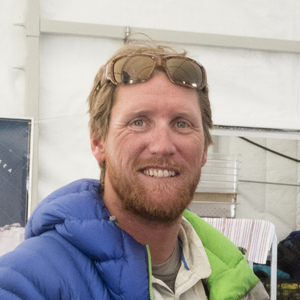 A man with short light brown hair and a beard, wearing sunglasses on his head and a blue puffer jacket, smiles while standing indoors with a white background, ready for his next Antarctic Logistics and Expeditions trip to Antarctica.