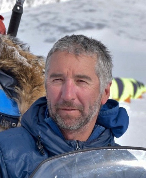 A man with short gray hair and a beard, wearing a blue winter jacket, stands outdoors in a snowy landscape near the South Pole. Fur-lined hoods and other Antarctic Logistics and Expeditions gear are visible behind him.