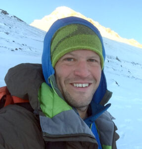 A man dressed in winter gear and a green beanie smiles outdoors on a snowy mountain slope in bright sunlight, capturing the spirit of Antarctic Logistics and Expeditions in Antarctica.