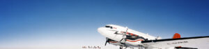 A small propeller plane with red and white markings sits on a snowy, flat landscape in Antarctica under a clear blue sky. Several people in cold-weather gear stand in the distance behind the plane, part of an Antarctic Logistics and Expeditions team.