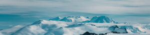 Snow-covered mountain peaks under a cloudy blue sky, with layers of white slopes and rugged ridges stretching across the Antarctic landscape near the South Pole.