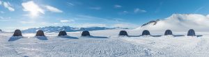 Seven dark tents are lined up on a snow-covered landscape in Antarctica, with mountains in the background under a bright, partly cloudy sky—a scene reminiscent of Antarctic Logistics and Expeditions near the South Pole.