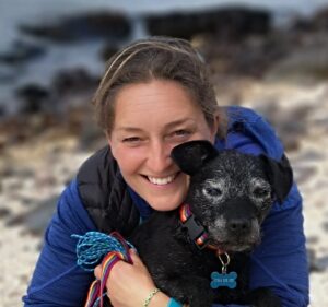 A smiling person with light brown hair, dressed in a blue top, hugs a small black dog with gray fur on its face. They are outdoors by a beach, reminiscent of Antarctica’s coastline, with blurry water and rocks in the background.