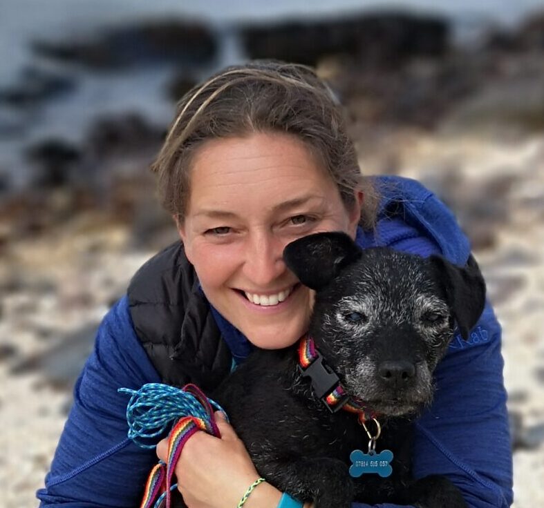A smiling person with light brown hair, dressed in a blue top, hugs a small black dog with gray fur on its face. They are outdoors by a beach, reminiscent of Antarctica’s coastline, with blurry water and rocks in the background.
