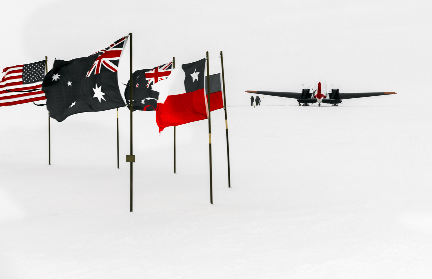 Several flags—including those of the United States, Australia, and New Zealand—stand in the snow at the South Pole, with a plane and two distant people set against Antarctica’s stark white landscape.