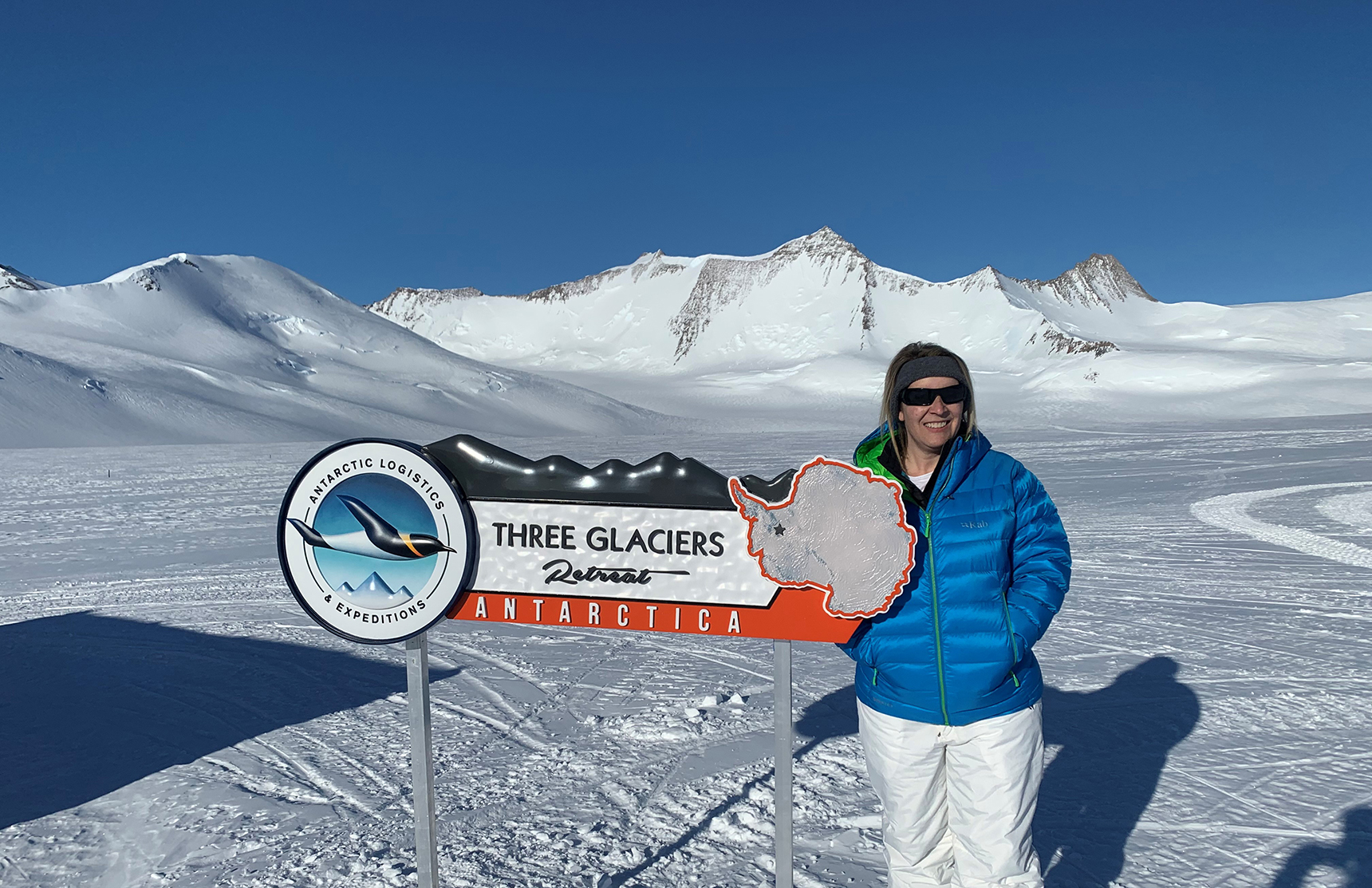 A person in a blue jacket and sunglasses stands beside a Three Glaciers Retreat Antarctica sign on a snowy landscape near distant mountains, under a clear blue sky, showcasing the spirit of Antarctic Logistics and Expeditions.