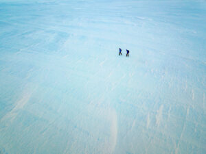 Two people walk side by side across a vast, snowy landscape in Antarctica. The minimal scene, with endless textured snow and ice, emphasizes the remoteness and scale of the South Pole environment.