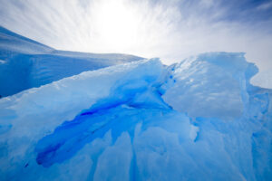 Bright sunlight shines over jagged, blue-tinted ice formations and snow-covered ground under a partly cloudy sky, capturing the cold, rugged beauty of Antarctica near the South Pole.