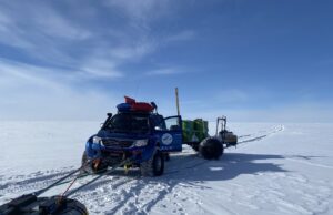A blue truck with oversized tires marked ALE (Antarctic Logistics and Expeditions) pulls equipment and supplies on sleds across a flat, snowy landscape in Antarctica under a clear, blue sky. Tire and sled tracks stretch across the pristine snow.