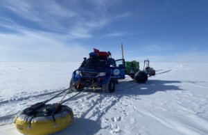 A blue off-road vehicle with gear on top pulls equipment on sleds and a yellow buoy across the vast, snowy expanse of Antarctica, supporting Antarctic Logistics and Expeditions under a clear blue sky.