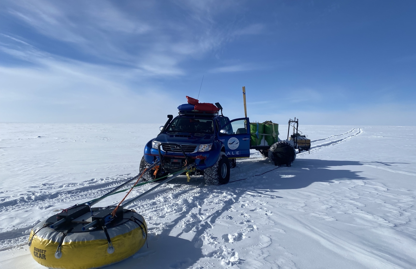 A blue off-road vehicle with gear on top pulls equipment on sleds and a yellow buoy across the vast, snowy expanse of Antarctica, supporting Antarctic Logistics and Expeditions under a clear blue sky.