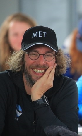 A smiling man with curly hair, glasses, and a black cap labeled MET rests his chin on his hand. Dressed in black and wearing a watch, he exudes the adventurous spirit of Antarctic Logistics and Expeditions, with blurred people in the background.