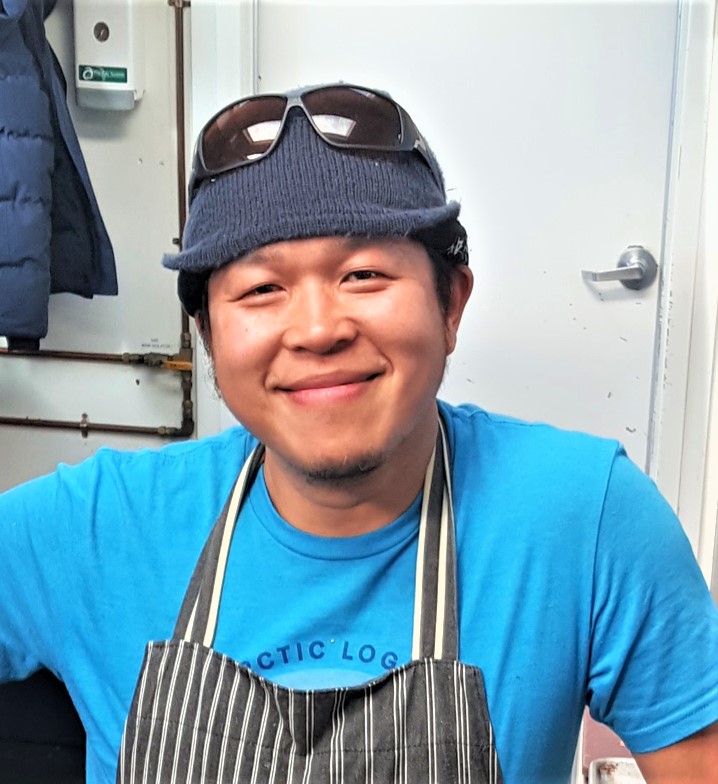 A smiling person wearing a blue shirt, striped apron, knit beanie, and sunglasses on their head stands indoors near a white door and wall hooks—ready for another busy day with Antarctic Logistics and Expeditions.