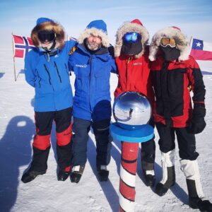 Four people in heavy winter gear stand together on snow at the South Pole next to a reflective sphere atop a striped pole, with flags of Norway and Chile visible in the background under a clear Antarctic sky.