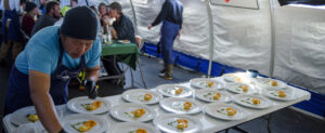A chef arranges plated dishes on a table inside a tent at the South Pole, each plate featuring elegant food portions. People sit and chat at tables in the background, creating a busy, communal dining atmosphere in remote Antarctica.