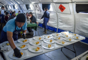 A chef wearing black gloves arranges food on white plates inside a large tent in Antarctica, while people enjoy their meals at tables in the background during an Antarctic Logistics and Expeditions event.