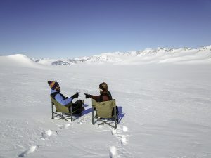 Two people sit in camping chairs on a snowy landscape, clinking glasses. Snow-covered mountains stretch across the horizon under a clear blue sky—an unforgettable moment at the South Pole with Antarctic Logistics and Expeditions.