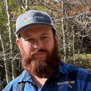 A bearded man in a plaid shirt and cap stands outdoors in sunlight, with leafless trees and dry branches behind him. His "Rufftech" cap and “Northern Tier” shirt evoke the rugged spirit of Antarctic Logistics and Expeditions in Antarctica.