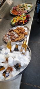 Three serving trays with assorted cheeses, fruits, meats, and cookies sit on a counter. In front, a large metal bowl filled with ice and Corona beers chills—perfect for an Antarctica-themed gathering or celebrating an Antarctic Logistics and Expeditions trip.