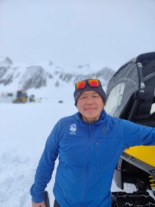 A person in a blue jacket and beanie, with reflective sunglasses on their head, stands in snowy terrain near a black and yellow vehicle. Snow-covered mountains and buildings evoke an Antarctic Logistics and Expeditions scene.