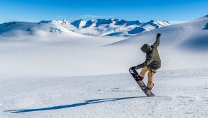 A snowboarder in a green jacket and tan pants performs a trick on a snowy slope in Antarctica, surrounded by snow-covered mountains under a clear blue sky, capturing the spirit of Antarctic Logistics and Expeditions.